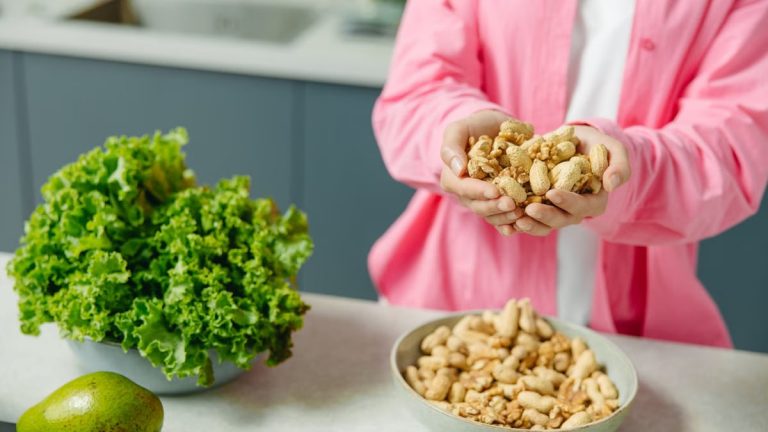 Hands presenting peanuts with a bowl, lettuce, avocado, and tomato nearby on kitchen counter.